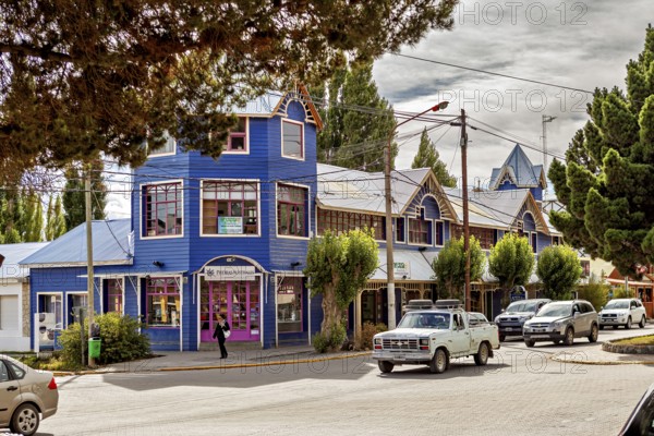 A blue, two-storey building in a busy street with parked cars and trees, street scene in El Chalten in Patagonia Argentina