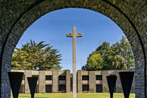 A stone cross stands in the centre of an arched passageway with a view of modern concrete architecture under a clear sky, German soldiers' cemetery from the Second World War near Huisnes-sur-Mer in Normandy, France