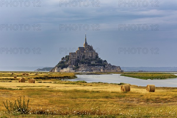 A castle island with ecclesiastical architecture in a rural landscape under a cloudy sky, Mont Saint Michel in Normandy, France