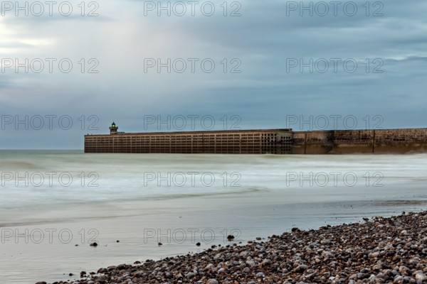 A breakwater juts into the sea, surrounded by a pebble beach and a cloudy sky, The pier and beach of Dieppe in Normandy, France