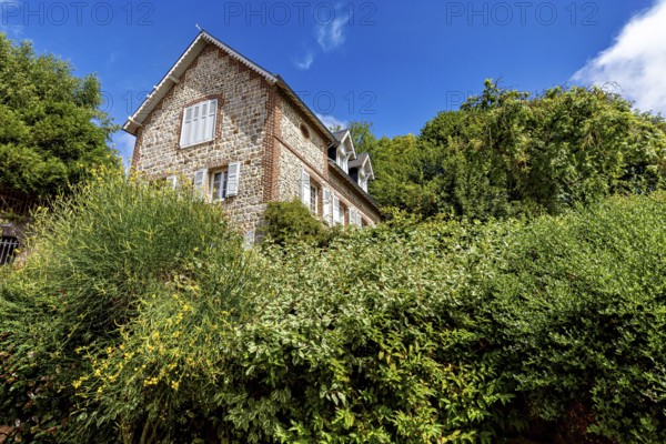 Single brick house behind green bushes and trees under a clear blue sky, The historic mill village of Veules Les Roses in Normandy, France