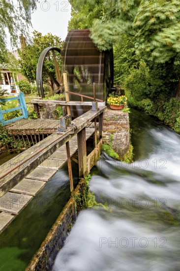 Old waterwheel next to a rushing river, surrounded by lush greenery and wooden bridges, the historic mill village of Veules Les Roses in Normandy, France