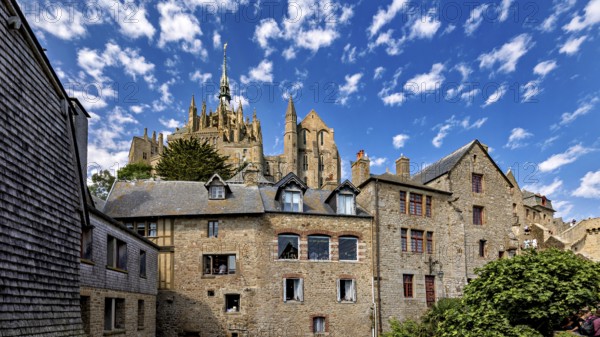 Historic stone architecture with a view of a castle under a blue sky with decorative clouds, Mont Saint Michel in Normandy, France
