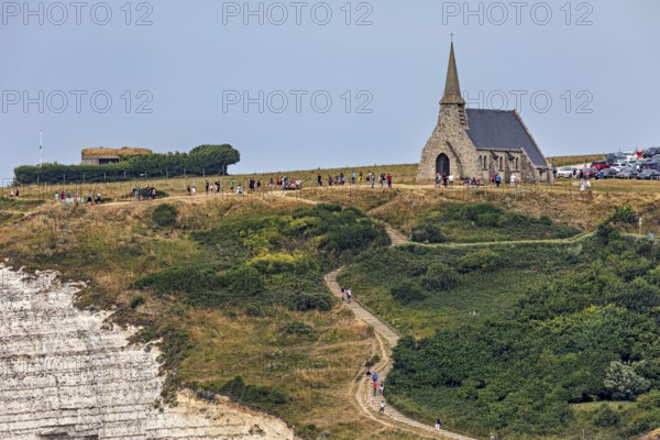 A small church stands on grassy cliffs with a path and walkers, under a blue sky, The church of Etretat in Normandy, France