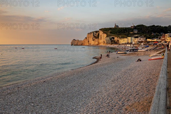 Beach with pebbles and boats, surrounded by rocks and a village, in the warm light of the sunset, The cliffs and alabaster coast of Etretat in Normandy France