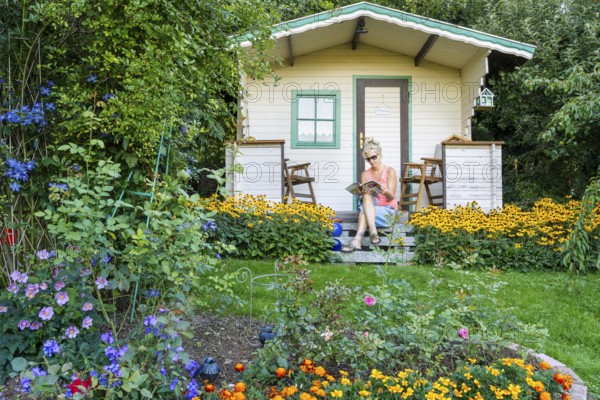 A woman sits relaxed on the small veranda of a garden shed and reads. Blooming summer flowers in the garden. Germany