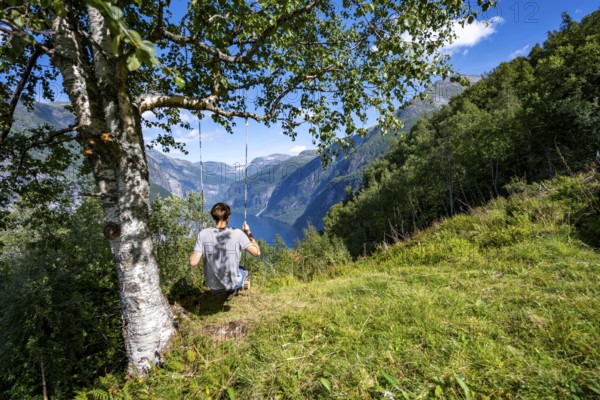 Young man swinging on a tree, view of idyllic fjord landscape of the Geirangerfjord at Blomberg Gård, near Geiranger, Møre og Romsdal, Norway