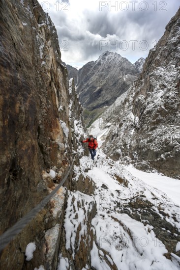 Mountaineer on a rope-secured path in a steep rocky mountain landscape with fresh snow in summer, behind mountain peaks Großer Friedrichskopf and Georgskopf, ascent to Hornscharte, Schobergruppe, Hohe Tauern National Park, Carinthia, Austria