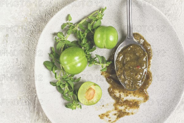 A spoon with tkemali sauce, on a plate, with cherry plum and mint, top view, no people