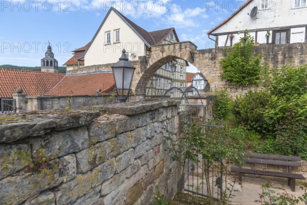 Former stone gate, town gate of the medieval town wall, Bad Sooden-Allendorf, district Allendorf, half-timbered houses, tower of St. Crucis church, bench, lamp, bushes, blue sky, clouds, Werra-Meissner district, Hesse, Germany