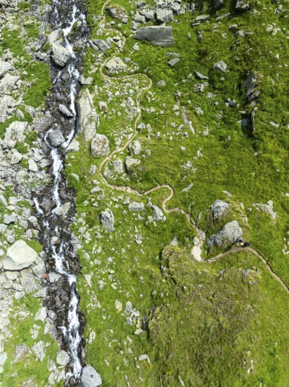 Hiking trail winds through mountain meadows interspersed with stones next to a mountain stream, from above, aerial view, Valais, Western Alps, Switzerland