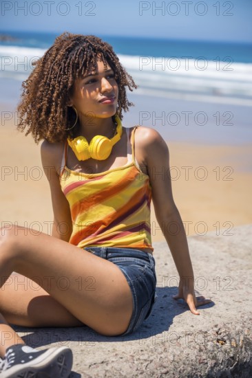 Young woman wearing headphones sitting on a rock at the beach, enjoying music and looking out at the ocean