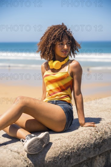 Young woman with curly hair and yellow headphones enjoys the beach view while sitting on a concrete wall