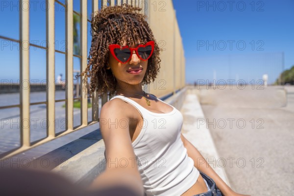 Fashionable young woman with curly hair enjoying a sunny day, taking a selfie by a stylish metal fence, radiating confidence and happiness