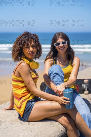 Two young women relaxing on beach wall, enjoying ocean view with skateboard and reusable coffee cup, embodying carefree summer vibes