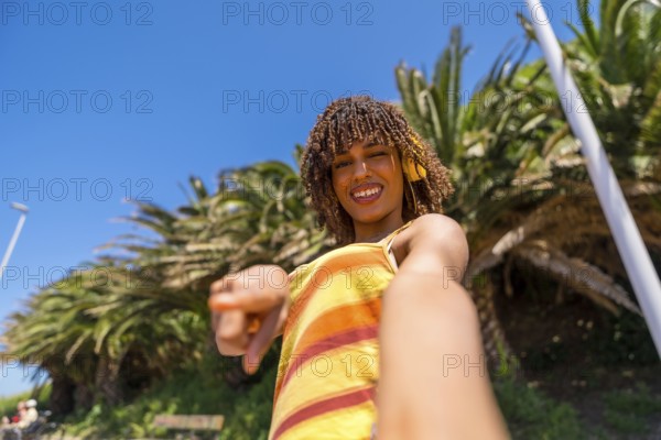 Carefree young woman with yellow headphones enjoying music, pointing finger and taking selfie in a tropical park