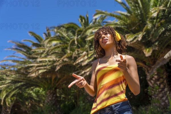 Carefree young woman with headphones enjoying music and pointing while standing under palm trees on a sunny summer day