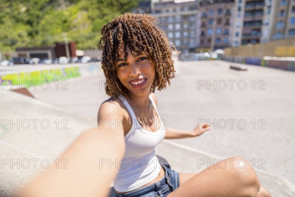 Smiling young woman with curly hair enjoying a sunny day at a skate park while taking a selfie portrait, capturing her vibrant urban lifestyle