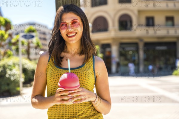 Happy young woman smiling brightly while enjoying a refreshing cold drink on a sunny summer day in the vibrant city streets