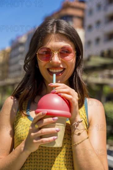 Happy young woman wearing stylish sunglasses, savoring a refreshing iced drink while enjoying a sunny day outdoors in the city