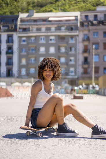 Young woman with curly hair sitting on her skateboard in an urban setting, enjoying a sunny day