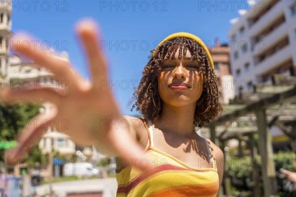 Stylish young woman extending her hand towards the camera in a vibrant city setting, enjoying a sunny summer day