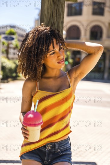 Stylish young woman with curly hair enjoys a cold beverage on a sunny summer day, exuding a carefree and vibrant spirit
