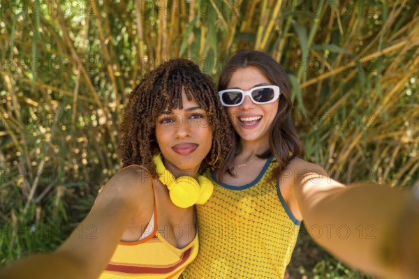 Two smiling girls taking a selfie in front of bamboo plants, wearing yellow clothes and headphones and sunglasses