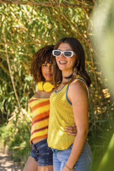 Two young women with headphones and sunglasses are embracing and smiling in a bamboo forest, enjoying a sunny day