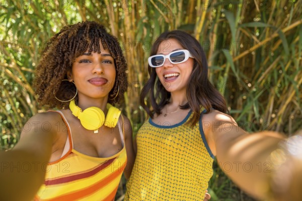 Two cheerful young women with colorful clothes and headphones taking a selfie in front of a bamboo forest