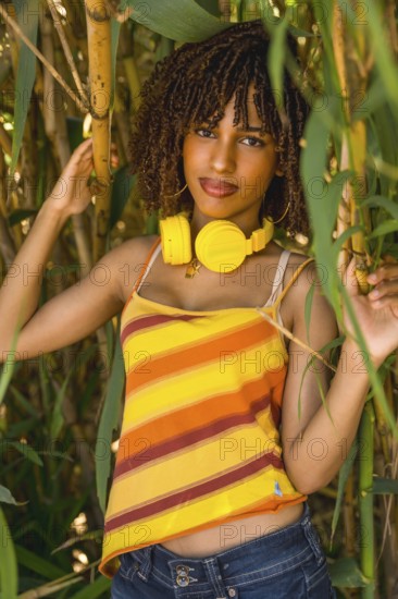 Young woman with yellow headphones enjoying music in a bamboo forest, wearing a colorful striped top and jeans
