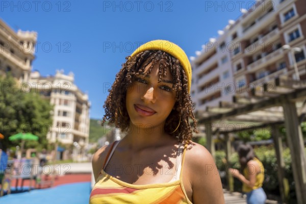 Portrait of a fashionable young woman with curly hair and yellow beanie enjoying a sunny day in an urban park