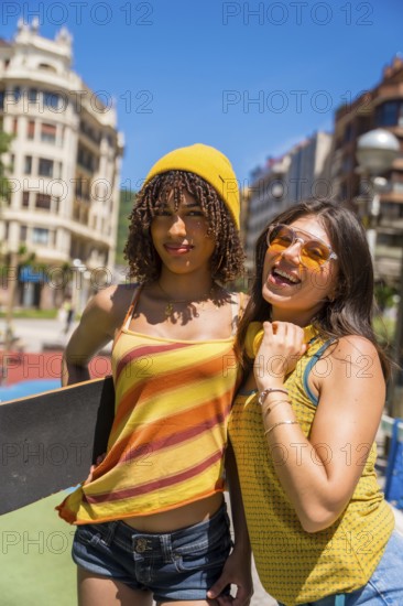 Two female friends enjoying a sunny day in the city, showcasing their youthful energy and urban style with skateboard and headphones