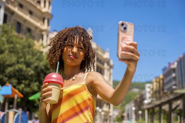 Stylish young woman captures a selfie while holding a refreshing beverage, enjoying a sunny day in a vibrant urban setting