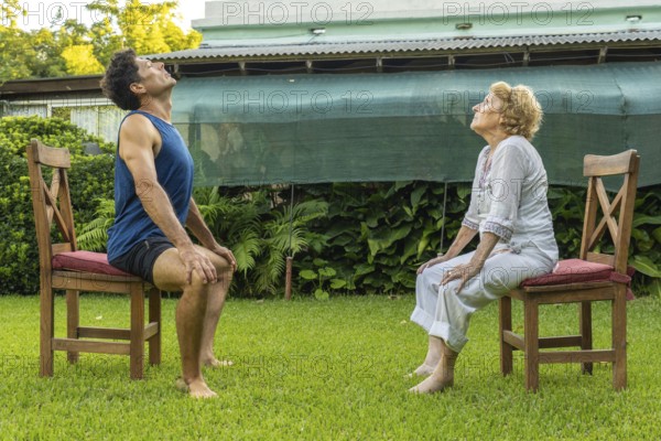 Senior woman and personal trainer engaging in seated exercises on chairs in a serene garden, supporting healthy aging and enhancing mobility through outdoor fitness activities