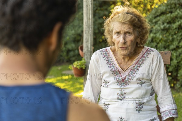 Elderly woman sitting comfortably on a wooden chair in a garden, engaging in a heartfelt conversation with her caregiver during a home visit, savoring the sunny day and fresh air
