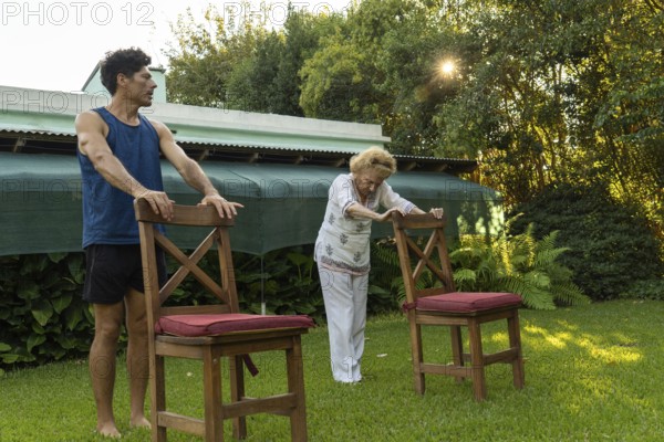 Senior woman practicing physical therapy exercises with the help of her personal trainer, using chairs as support in a backyard setting, promoting health and mobility in old age