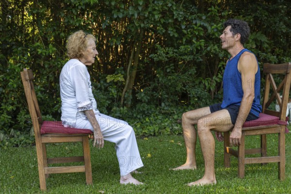 Senior woman and her personal trainer practicing rehabilitation exercises sitting on chairs in a garden, promoting healthy aging and active lifestyle