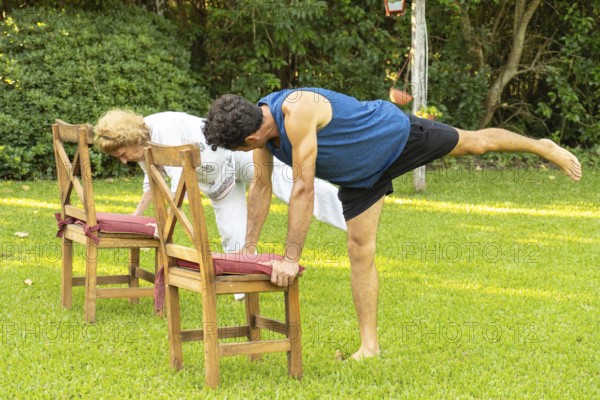 Senior man and woman practicing yoga in garden, doing balancing pose using chairs for support, promoting healthy lifestyle and fitness in old age