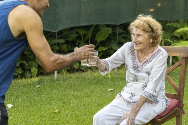 Young male nurse is offering a glass of fresh water to a smiling elderly woman sitting on a wooden chair in a garden, showing care and support for the elderly