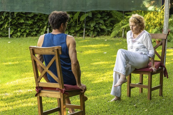 Senior woman and her physiotherapist are sitting on chairs in a garden, practicing rehabilitation exercises to improve mobility and well-being