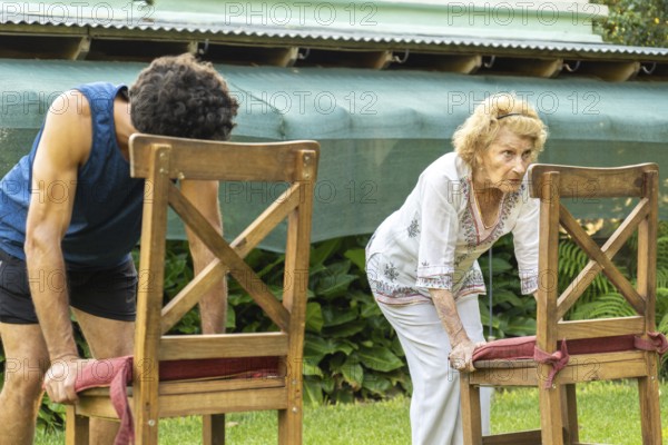 Senior woman and her personal trainer bending over while gripping chairs, engaging in back muscle exercises in a lush garden setting, promoting fitness and healthy aging