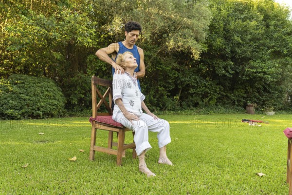 Physiotherapist is giving a shoulder massage to a senior woman sitting on a chair in a peaceful garden setting, promoting relaxation and well-being
