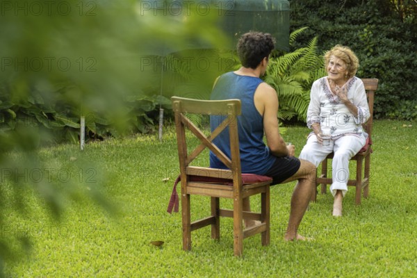 Young male nurse assisting an elderly woman during a home visit, engaging in conversation while sitting on chairs in a lush green garden, promoting well-being and personalized care