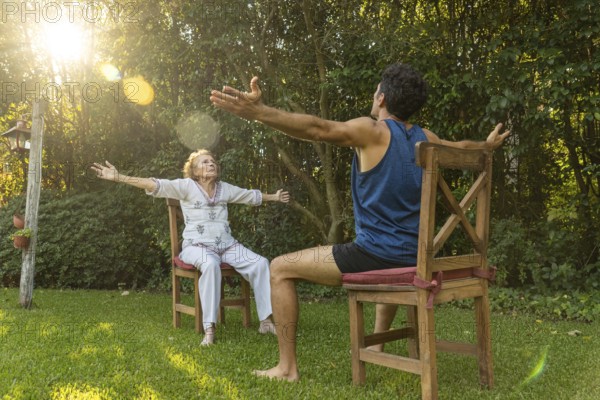 Senior woman practicing seated exercises with the assistance of a physiotherapist in a green backyard, promoting health and mobility in old age