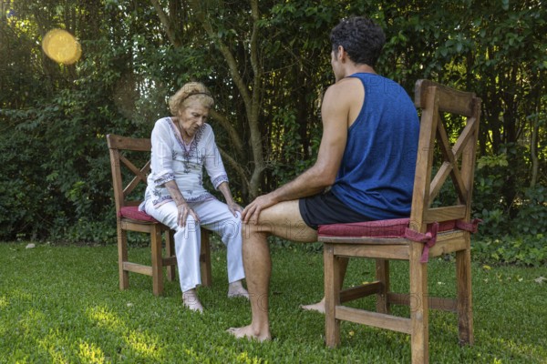 Senior woman experiencing knee pain receiving assistance from a physiotherapist during an outdoor therapy session in a peaceful garden setting, promoting health and well-being in old age