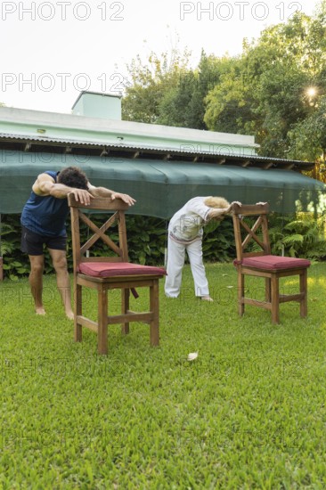 Senior woman and her physiotherapist are bending forward, holding onto chairs in a garden, performing stretching exercises to improve flexibility and mobility