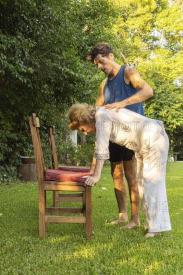 Physiotherapist is assisting a senior woman performing bending exercises with the help of a chair in a lush green garden, promoting mobility and well-being