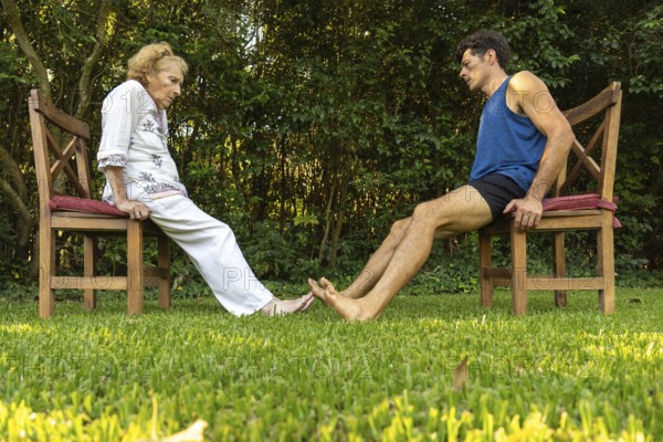 Elderly woman and her personal trainer practicing leg strengthening exercises using chairs in a garden, promoting healthy aging and mobility