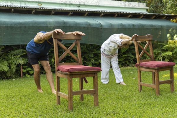 Elderly man and woman practicing stretching exercises with chairs on a grassy lawn in a serene garden setting, promoting health and flexibility in old age